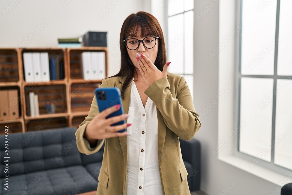 Sticker young brunette woman working at the office with smartphone covering mouth with hand, shocked and afr