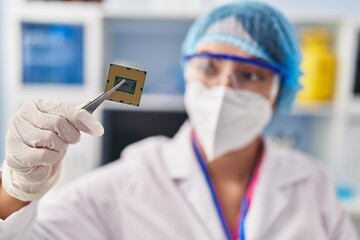 Young beautiful hispanic woman scientist wearing medical mask holding cpu processor chip at pharmacy