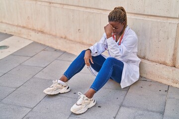 African american woman doctor stressed sitting on floor at street