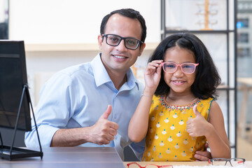 Indian cute little daughter and father are choosing appropriate eyeglasses for children in...