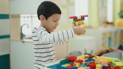 Adorable hispanic boy playing with construction blocks sitting on table at kindergarten