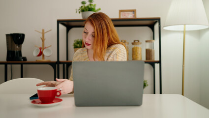 Young redhead woman using laptop drinking coffee at home
