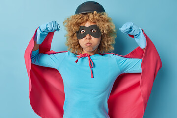 Surprised woman wears helmet eyemask and cloak raises arms shows biceps blows cheeks stares bugged...