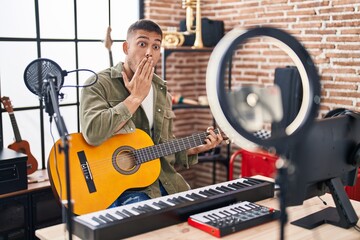 Young hispanic man doing online guitar tutorial covering mouth with hand, shocked and afraid for...