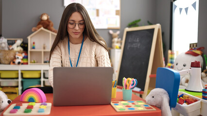 Young beautiful hispanic woman preschool teacher using laptop sitting on table at kindergarten