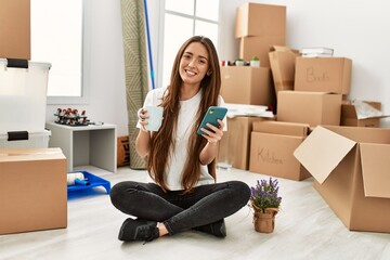 Young hispanic woman using smartphone drinking coffee sitting on floor at new home
