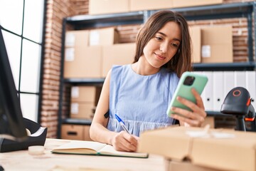 Young beautiful hispanic woman ecommerce business worker using smartphone writing on notebook at office