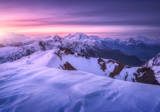 Snow covered mountains and colorful purple sky with clouds at sunset in winter. Beautiful wintry landscape with snowy rocks and hills at twilight. Scenery with alps at frosty evening. Alpine mountains