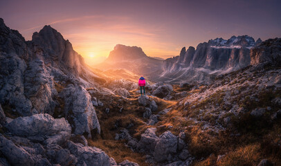 Fototapeta premium Woman on the mountain trail and stones at sunset in autumn. Hiking in Dolomites, Italy. Girl on the path and high rocks at twilight in fall. Colorful landscape with cliffs, grass, golden sky. Nature