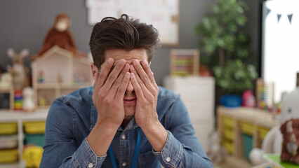 Young hispanic man preschool teacher stressed sitting on table at kindergarten