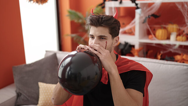 Young hispanic man wearing devil costume inflating balloon at home