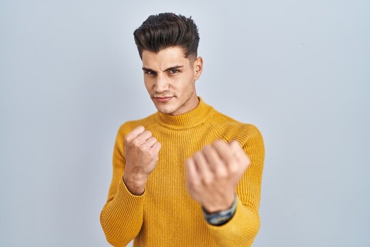 Young hispanic man standing over blue background ready to fight with fist defense gesture, angry and upset face, afraid of problem
