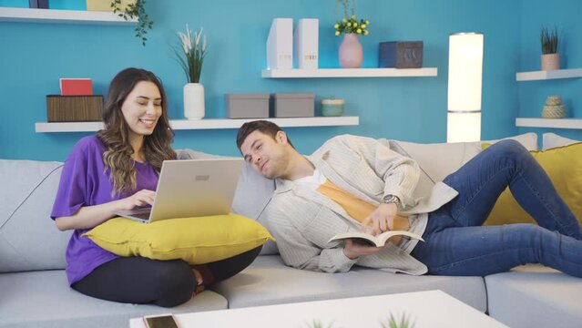Happy couple sitting on sofa at home having a pleasant and intimate time.
Happy couple having fun together at home. The man reading a book is looking at his wife working on the laptop, they are chatti