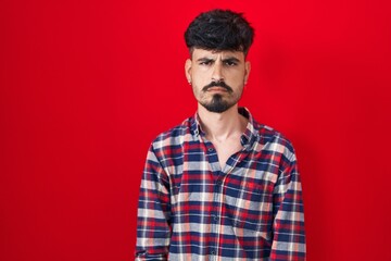 Young hispanic man with beard standing over red background skeptic and nervous, frowning upset because of problem. negative person.