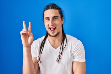 Hispanic man with long hair standing over blue background smiling with happy face winking at the...