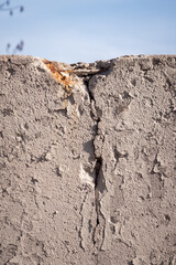 Close up of a crack in a concrete masonry wall with rust and rebar starting to show through with layers of beige or tan colored off white peeling paint and blue sky beyond.