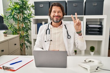 Handsome middle age doctor man working at the clinic showing and pointing up with fingers number two while smiling confident and happy.