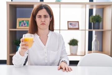 Brunette woman drinking glass of orange juice skeptic and nervous, frowning upset because of problem. negative person.