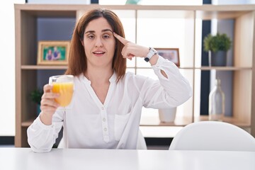 Brunette woman drinking glass of orange juice smiling pointing to head with one finger, great idea or thought, good memory