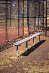 Close up of an empty aluminum metal baseball bench in a grass and dirt dugout with black chain link fence and muddy red colored infield beyond.
