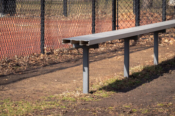 Close up of an empty aluminum metal baseball bench in a grass and dirt dugout with black chain link...