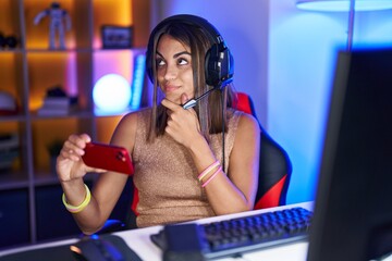 Young hispanic woman playing video games with smartphone serious face thinking about question with hand on chin, thoughtful about confusing idea