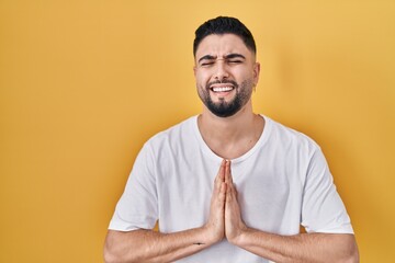 Young handsome man wearing casual t shirt over yellow background begging and praying with hands together with hope expression on face very emotional and worried. begging.