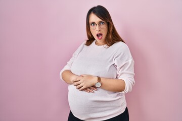 Pregnant woman standing over pink background in shock face, looking skeptical and sarcastic, surprised with open mouth