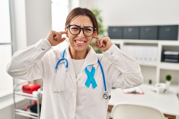 Young brunette doctor woman wearing stethoscope at the clinic covering ears with fingers with annoyed expression for the noise of loud music. deaf concept.
