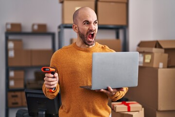 Young bald man with beard working at small business ecommerce with laptop angry and mad screaming frustrated and furious, shouting with anger. rage and aggressive concept.