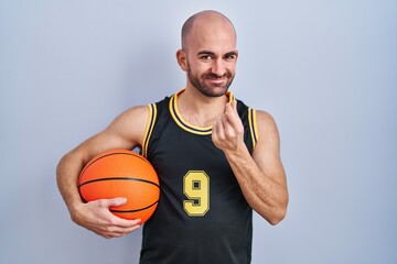 Young bald man with beard wearing basketball uniform holding ball doing money gesture with hands,...