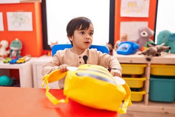 Adorable hispanic boy student opening backpack sitting on table at kindergarten