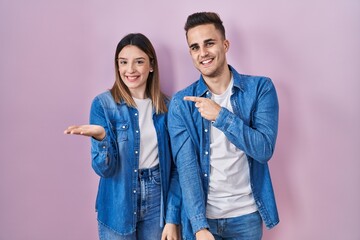 Young hispanic couple standing over pink background amazed and smiling to the camera while presenting with hand and pointing with finger.