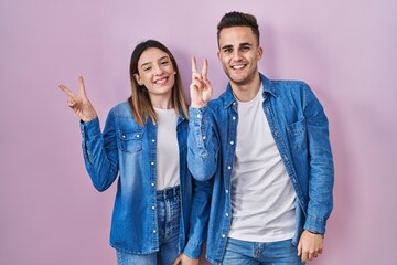 Fototapeta premium Young hispanic couple standing over pink background smiling with happy face winking at the camera doing victory sign with fingers. number two.