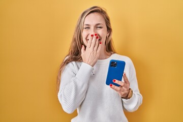 Young blonde woman using smartphone typing message laughing and embarrassed giggle covering mouth...