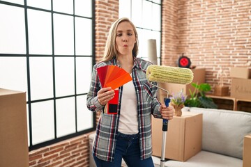 Young blonde woman holding roller painter and paint samples at new home making fish face with mouth and squinting eyes, crazy and comical.