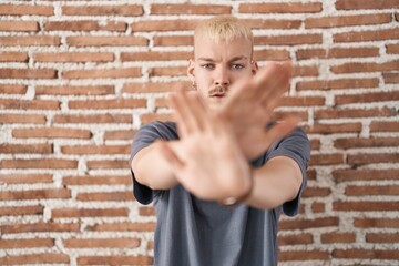 Young caucasian man standing over bricks wall rejection expression crossing arms and palms doing negative sign, angry face