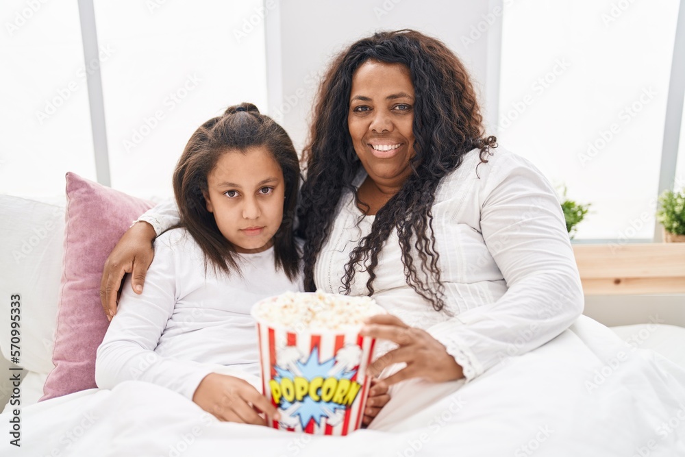 Poster mother and young daughter eating popcorn in the bed relaxed with serious expression on face. simple 