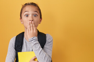 Little caucasian boy wearing student backpack and holding book covering mouth with hand, shocked...