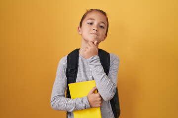Little caucasian boy wearing student backpack and holding book serious face thinking about question with hand on chin, thoughtful about confusing idea