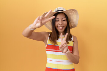 Middle age chinese woman wearing summer hat over yellow background smiling with tongue out showing...