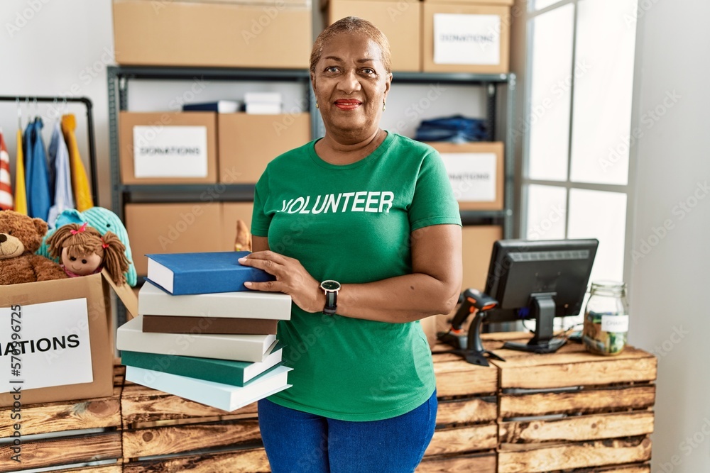 Canvas Prints Senior african american woman wearing volunteer uniform holding books at charity center