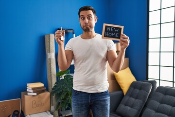 Young hispanic man with beard holding keys of new home puffing cheeks with funny face. mouth...