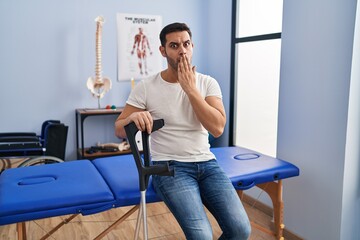 Young hispanic man with beard wearing crutches at rehabilitation clinic covering mouth with hand,...