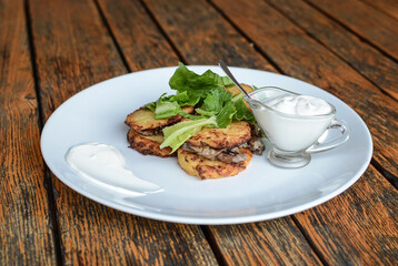 Pancakes from grated potatoes with mushrooms and sour cream on a white plate on a wooden background.	