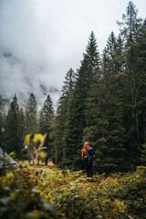 Obraz premium A hiker with orange raincoat is exploring the surroundings of an isolated mountain hut in the forest of Val di Genova, during a rainy and foggy autumnal day, Trentino Alto Adige, Northern Italy