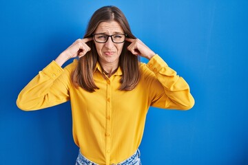 Young woman wearing glasses standing over blue background covering ears with fingers with annoyed expression for the noise of loud music. deaf concept.