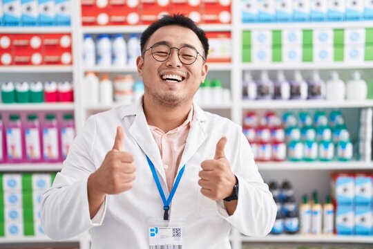Chinese young man working at pharmacy drugstore success sign doing positive gesture with hand, thumbs up smiling and happy. cheerful expression and winner gesture.