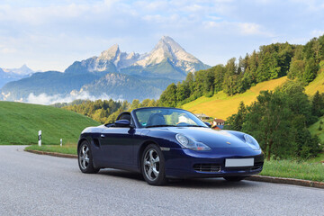 Berchtesgaden, Germany - July 25, 2021: Blue roadster Porsche Boxster 986 with mountain Watzmann and fog panorama. The car is a mid-engine two-seater sports car manufactured by Porsche.