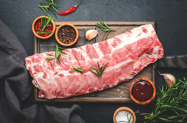 Raw pork ribs with rosemary and spices on rustic wooden cutting board prepared for cooking on black kitchen table background, top view